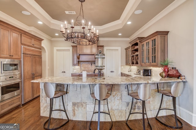 kitchen with kitchen peninsula, a kitchen breakfast bar, light stone countertops, a tray ceiling, and built in appliances