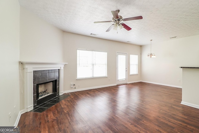 unfurnished living room with ceiling fan with notable chandelier, a tile fireplace, a textured ceiling, and dark wood-type flooring