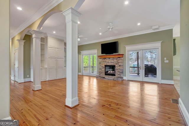 unfurnished living room with a stone fireplace, crown molding, ceiling fan, light wood-type flooring, and decorative columns