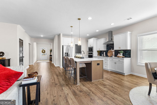 kitchen with a center island with sink, appliances with stainless steel finishes, pendant lighting, wall chimney exhaust hood, and white cabinets