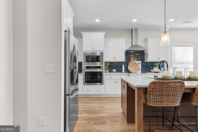 kitchen featuring stainless steel appliances, wall chimney range hood, hanging light fixtures, and white cabinetry