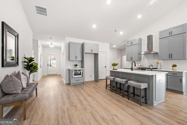 kitchen with a kitchen breakfast bar, wall chimney exhaust hood, a kitchen island with sink, light hardwood / wood-style flooring, and gray cabinets