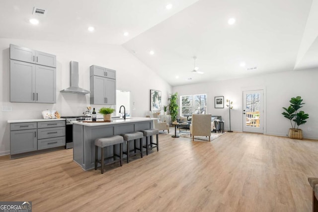 kitchen with gray cabinetry, wall chimney exhaust hood, light hardwood / wood-style flooring, a center island with sink, and stainless steel stove