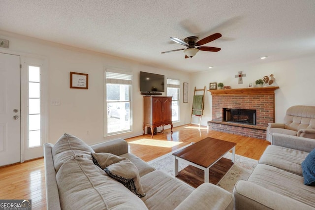 living room featuring a brick fireplace, light hardwood / wood-style flooring, ceiling fan, a textured ceiling, and a wealth of natural light