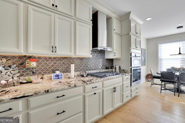 kitchen with wall chimney range hood, hanging light fixtures, light hardwood / wood-style floors, white cabinetry, and stainless steel appliances