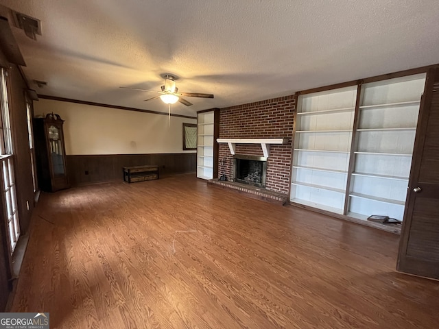 unfurnished living room with a textured ceiling, ceiling fan, wood-type flooring, a brick fireplace, and crown molding