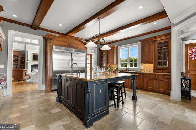 kitchen with light stone countertops, stainless steel built in refrigerator, pendant lighting, a center island with sink, and beamed ceiling