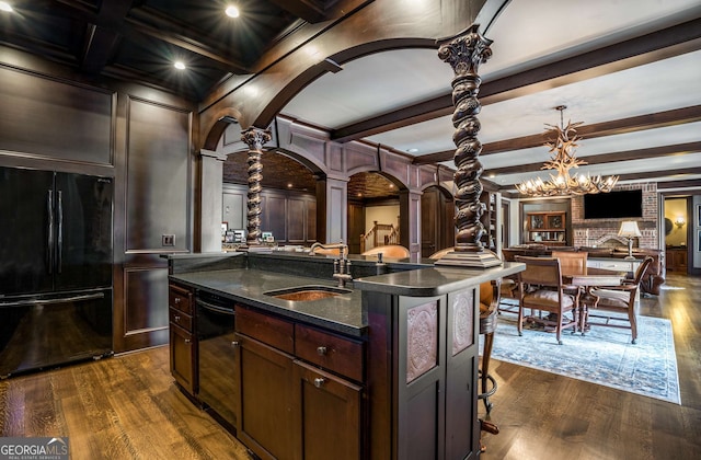 kitchen with decorative columns, sink, black appliances, beamed ceiling, and an island with sink