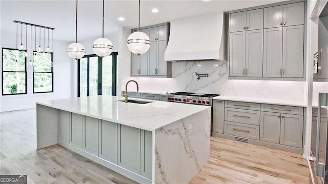 kitchen with custom exhaust hood, an island with sink, light stone counters, sink, and backsplash