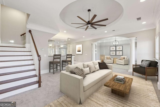 carpeted living room featuring ceiling fan with notable chandelier, ornamental molding, a tray ceiling, and decorative columns