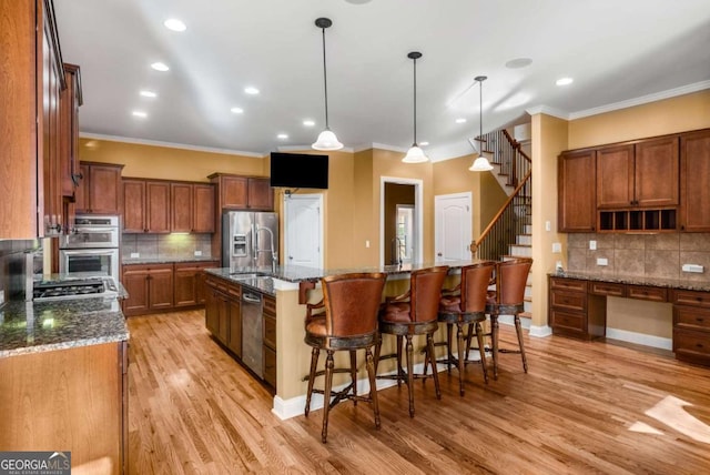 kitchen featuring dark stone countertops, light wood-type flooring, an island with sink, hanging light fixtures, and appliances with stainless steel finishes