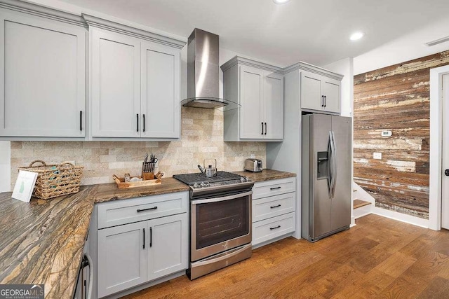 kitchen featuring wall chimney range hood, light hardwood / wood-style floors, wooden walls, gray cabinetry, and appliances with stainless steel finishes