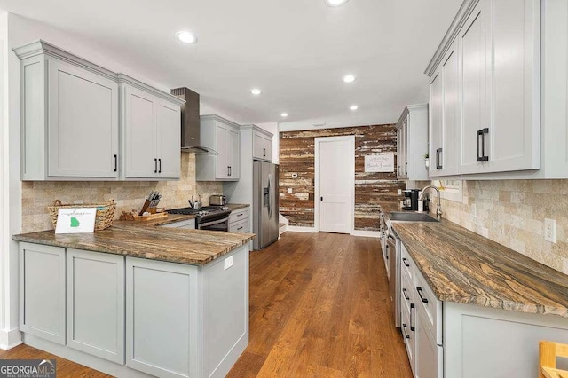 kitchen with dark wood finished floors, dark stone counters, wall chimney exhaust hood, stainless steel appliances, and a sink