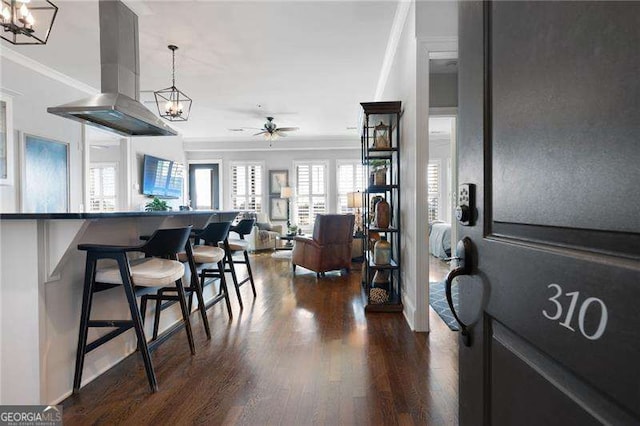 kitchen featuring a kitchen bar, ceiling fan, ornamental molding, island range hood, and dark wood-type flooring