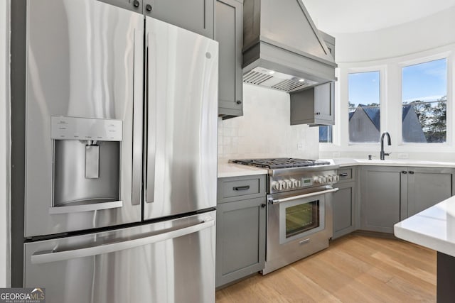 kitchen with custom exhaust hood, gray cabinets, stainless steel appliances, sink, and light hardwood / wood-style flooring