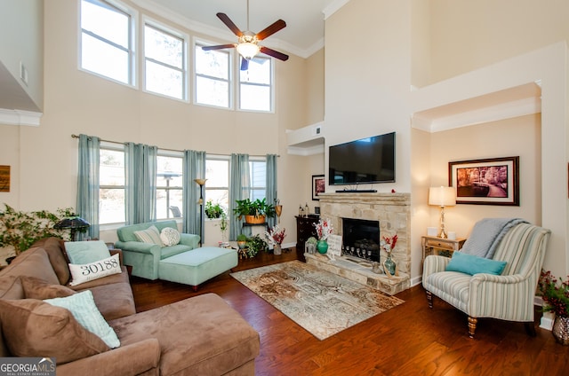 living room featuring ceiling fan, ornamental molding, dark hardwood / wood-style floors, a high ceiling, and a stone fireplace