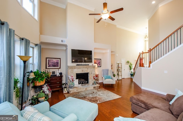 living room with dark wood-type flooring, a towering ceiling, crown molding, ceiling fan, and a stone fireplace