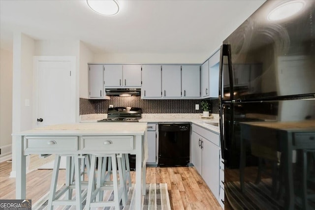 kitchen featuring light wood finished floors, under cabinet range hood, light countertops, black appliances, and backsplash