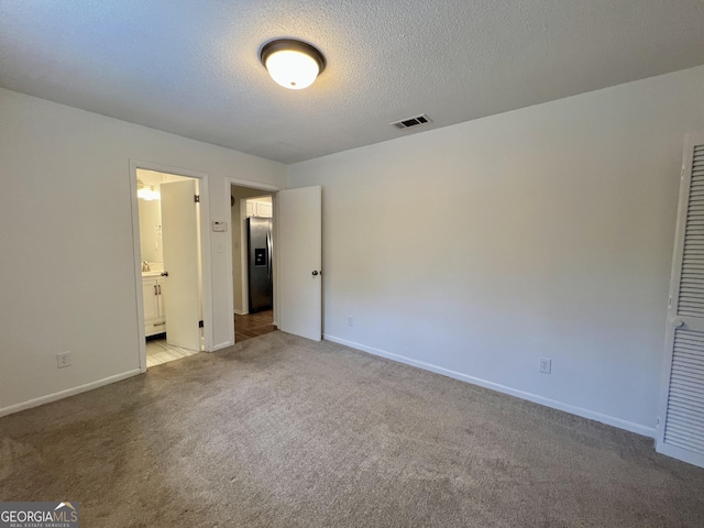 unfurnished bedroom with stainless steel refrigerator with ice dispenser, ensuite bath, a textured ceiling, and light colored carpet