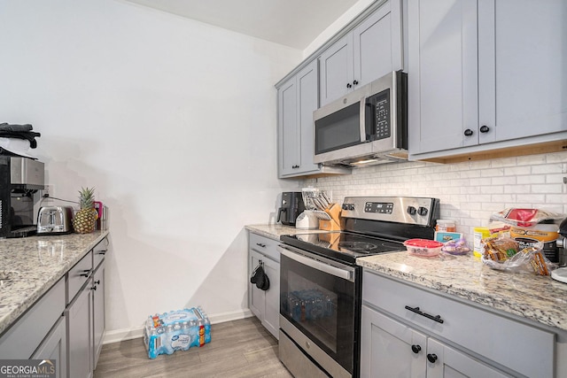 kitchen featuring gray cabinets, light hardwood / wood-style flooring, decorative backsplash, light stone countertops, and appliances with stainless steel finishes