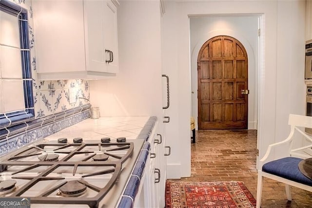 kitchen featuring brick floor, arched walkways, tasteful backsplash, tile counters, and white cabinets