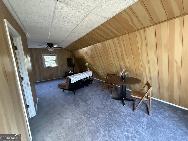 sitting room with wood walls, ceiling fan, vaulted ceiling, and dark colored carpet