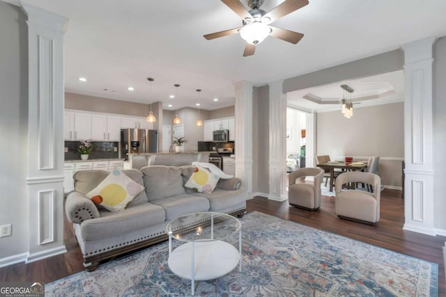 living room featuring ceiling fan, decorative columns, a tray ceiling, and dark wood-type flooring