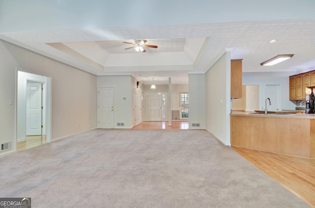 unfurnished living room with light carpet, sink, ornamental molding, ceiling fan, and a tray ceiling