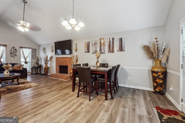 dining space with light hardwood / wood-style floors, a brick fireplace, vaulted ceiling, and ceiling fan with notable chandelier