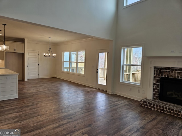 unfurnished living room with dark wood-type flooring, an inviting chandelier, crown molding, baseboards, and a brick fireplace