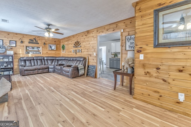 living room featuring ceiling fan, wood walls, light wood-type flooring, and a textured ceiling