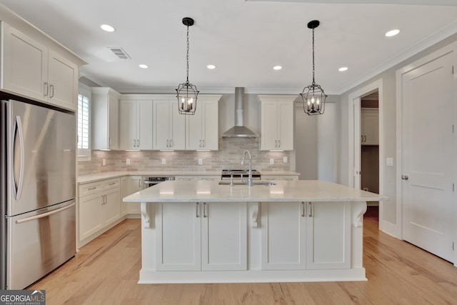 kitchen featuring stainless steel appliances, wall chimney exhaust hood, an island with sink, and hanging light fixtures