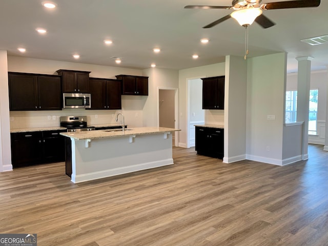 kitchen with appliances with stainless steel finishes, an island with sink, sink, light stone counters, and light wood-type flooring