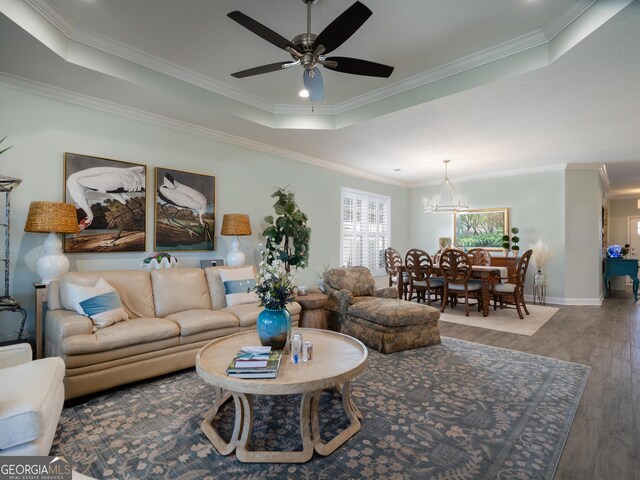 living room featuring wood-type flooring, ornamental molding, ceiling fan with notable chandelier, and a tray ceiling