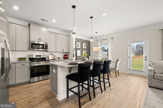 kitchen featuring decorative light fixtures, an island with sink, backsplash, gray cabinetry, and appliances with stainless steel finishes