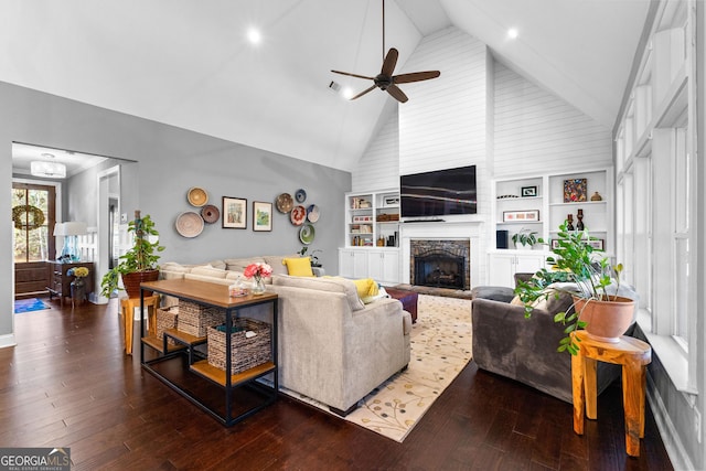 living room featuring dark wood-type flooring, built in shelves, high vaulted ceiling, ceiling fan, and a stone fireplace