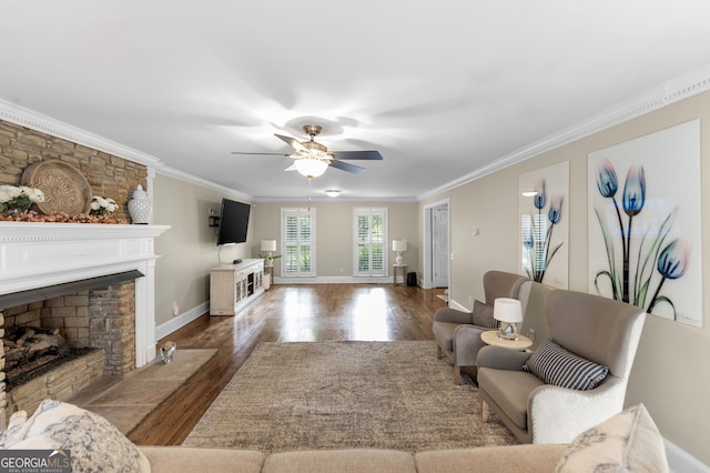 living room featuring dark wood-type flooring, ceiling fan, crown molding, and a stone fireplace