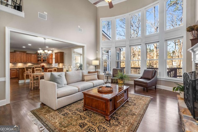 living room with a stone fireplace, ornamental molding, dark wood-type flooring, a high ceiling, and ceiling fan with notable chandelier