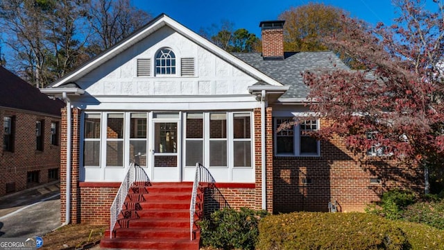 view of front of home with a shingled roof, brick siding, a sunroom, and a chimney