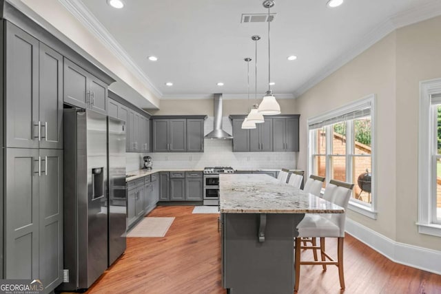 kitchen featuring a kitchen bar, hanging light fixtures, appliances with stainless steel finishes, gray cabinets, and wall chimney range hood