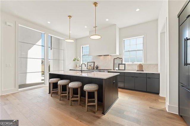 kitchen featuring hanging light fixtures, an island with sink, light wood-type flooring, white cabinets, and tasteful backsplash