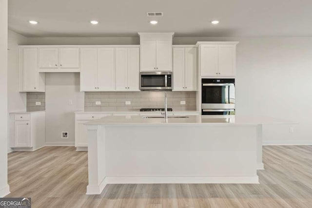 kitchen with stainless steel appliances, white cabinets, light hardwood / wood-style floors, decorative backsplash, and a center island with sink