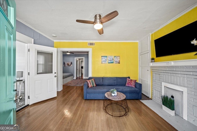 living room featuring a brick fireplace, wood-type flooring, ornamental molding, and ceiling fan