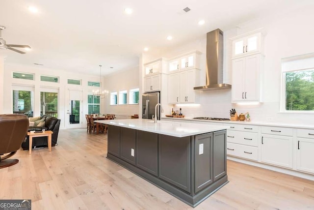 kitchen with wall chimney exhaust hood, hanging light fixtures, stainless steel fridge, a kitchen island with sink, and white cabinetry