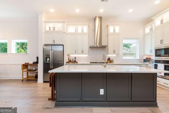 kitchen with stainless steel appliances, wall chimney range hood, an island with sink, and white cabinetry