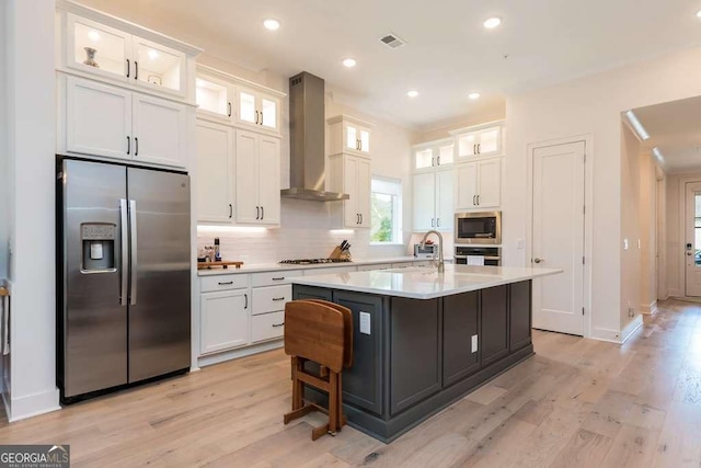 kitchen with stainless steel appliances, a kitchen island with sink, wall chimney range hood, and white cabinets