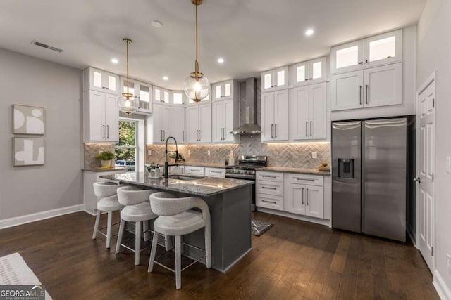 kitchen featuring white cabinetry, dark stone countertops, an island with sink, wall chimney range hood, and appliances with stainless steel finishes