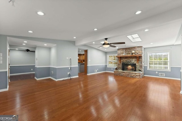 unfurnished living room with a stone fireplace, ceiling fan, vaulted ceiling with skylight, and hardwood / wood-style floors