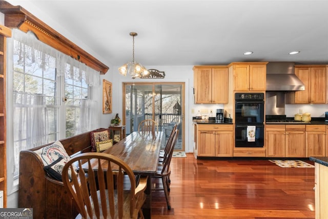 kitchen featuring black double oven, pendant lighting, dark hardwood / wood-style floors, wall chimney range hood, and an inviting chandelier