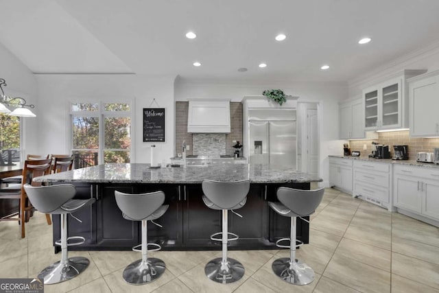 kitchen featuring white cabinetry, decorative backsplash, stainless steel built in fridge, light stone countertops, and a large island with sink